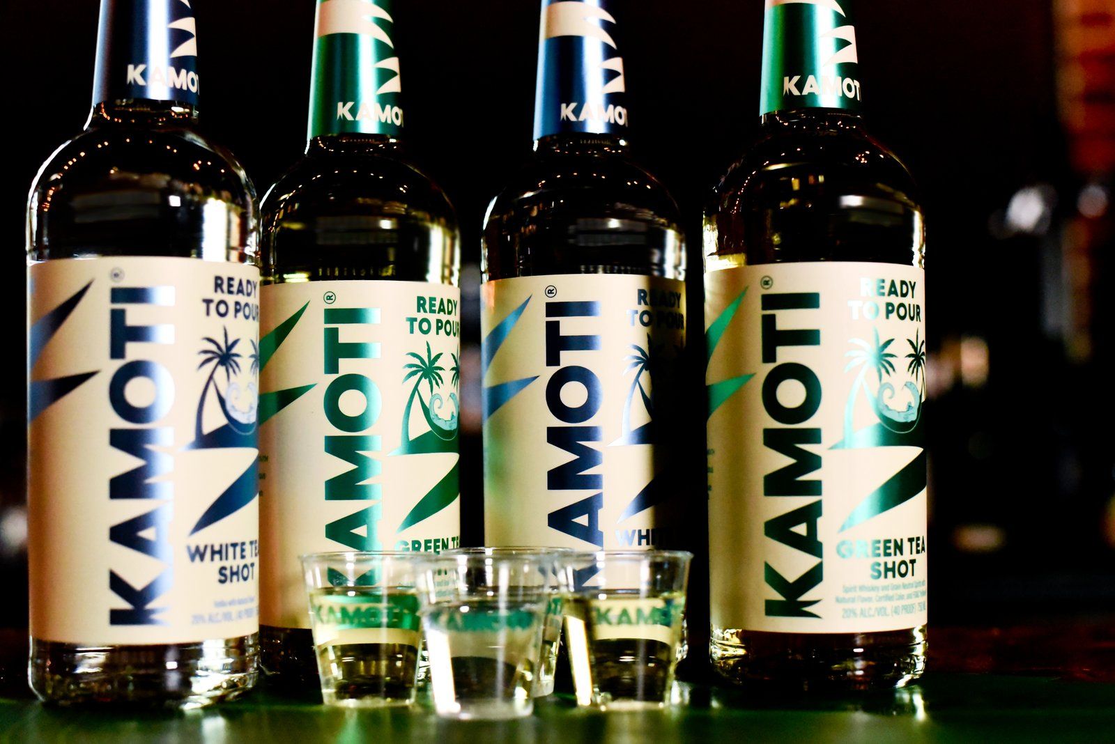 Four Kamoti white tea and green tea shot bottles behind three filled shot glasses on a bar