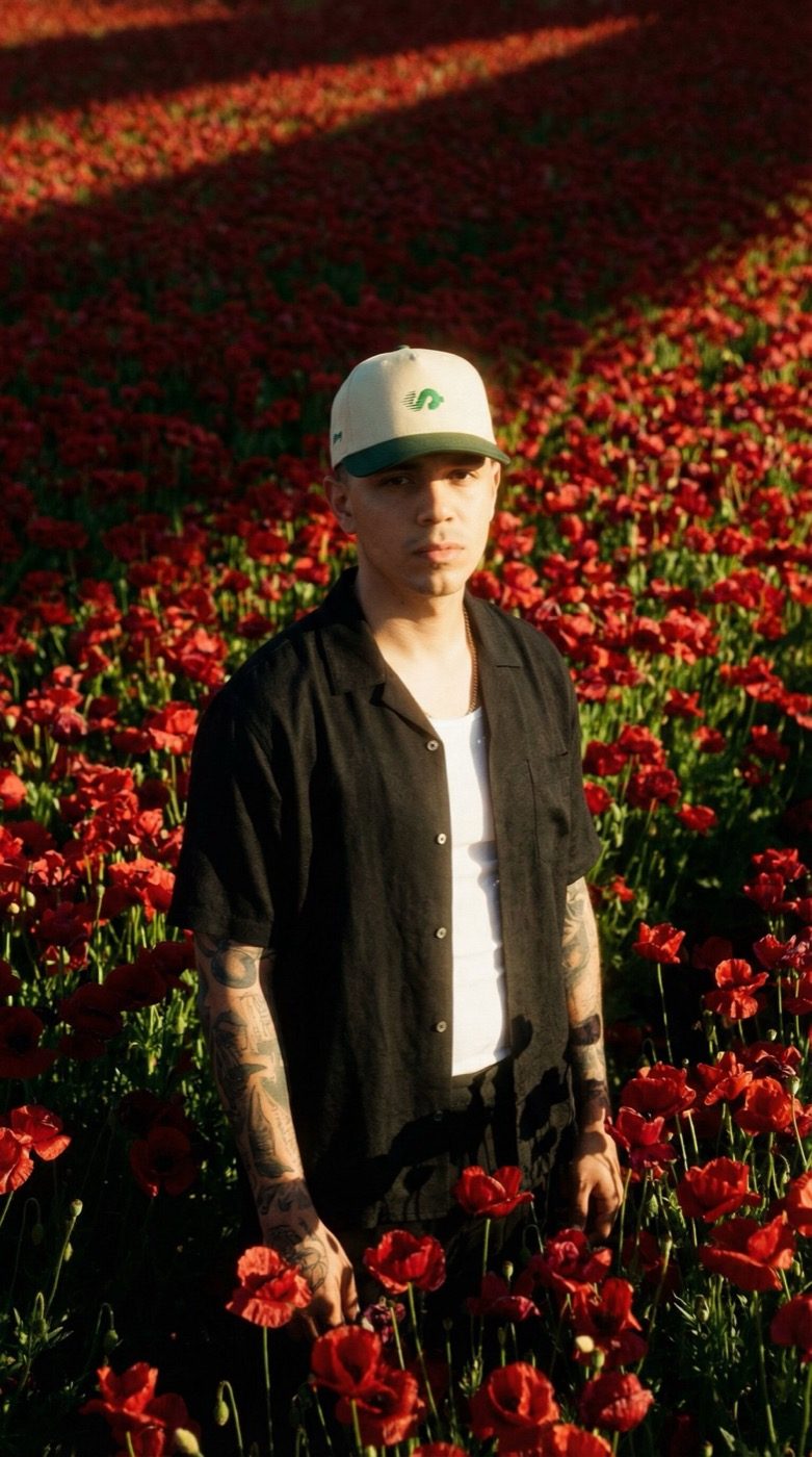 Product photograph: a model in a cream and green Sidemoney cap and a black shirt stands in a field of red poppies