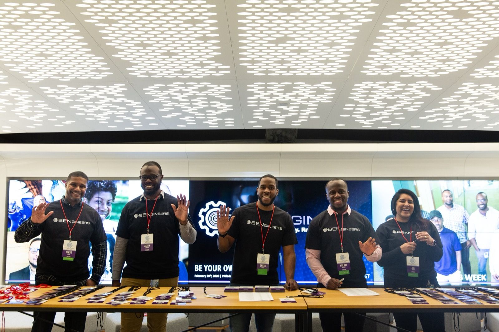 Five BENgineers volunteers in black BENgineers tees standing behind a conference table, with the group wordmark on the screens behind them